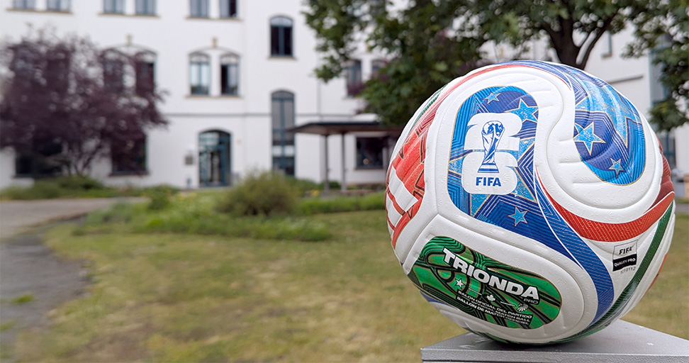 A close-up of a colorful FIFA World Cup soccer ball resting on a pedestal, with a building and trees in the background. The ball features a logo and vivid designs in red, blue, and green.