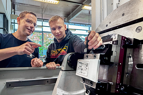 Two individuals are engaged in a discussion in a workshop setting. One person, wearing glasses, points to a machine component while the other looks on attentively. They appear to be discussing a technical procedure related to the machinery they are working with.