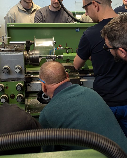 A group of individuals is observing a person working on a lathe machine. The focus is on the machine's operation, with multiple people engaged in the process. Tools and equipment are present in the environment, indicating a manufacturing or workshop setting.