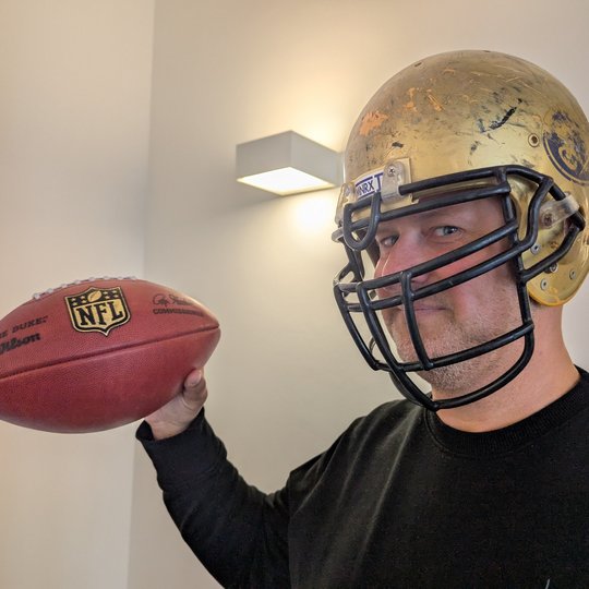 Ein Mann trägt einen goldenen Footballhelm und hält einen Football mit dem NFL-Logo in der Hand. Er schaut direkt in die Kamera und trägt ein schwarzes Sweatshirt. Der Hintergrund ist hell und unförmig.