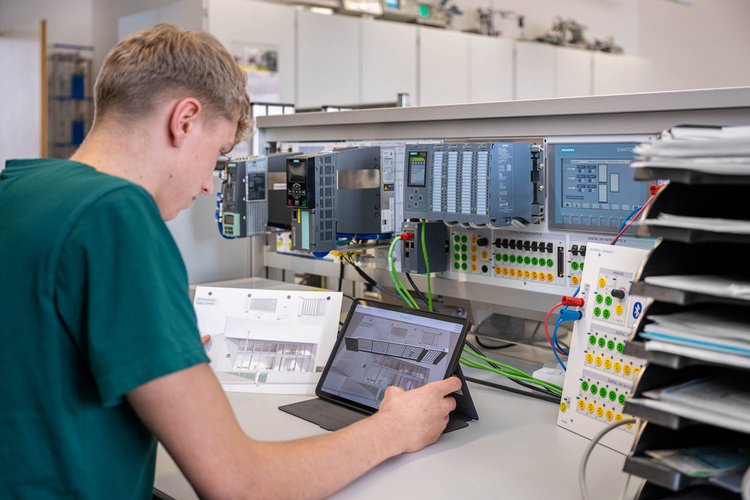 A young man is seated at a workbench, studying a tablet while referencing a paper document. Various electronic components and control panels are arranged behind him, indicating a technical or engineering environment. The setting suggests hands-on learning or work in automation or programming.