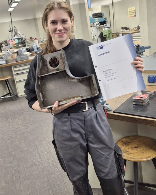 A young woman stands in a workshop, smiling while holding a metal part and a document titled "Zeugnisse." She is wearing a black shirt and gray pants, with tools and machinery visible in the background.