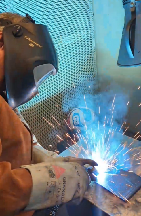 A welder, wearing protective gear and a helmet, is focused on welding a piece of metal. Bright sparks and blue light emanate from the weld area, creating a hazy atmosphere filled with smoke.