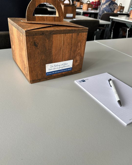 Eine holzfarbene Box mit einem Griff steht auf einem Tisch. An der Box befindet sich ein Aufkleber mit dem Text: "Von Bildung und Kleinen kann man viel lernen." Neben der Box liegt ein Notizblock mit einem Stift.