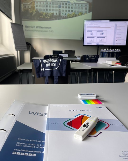 Ein Tisch in einem Seminarraum mit Schulungsunterlagen zum Thema Arbeitsrecht. Ein Stift und farbige Marker liegen neben den Unterlagen. Im Hintergrund ist eine Präsentation mit der Begrüßung „Herzlich Willkommen“ und dem Logo der IHK sichtbar.