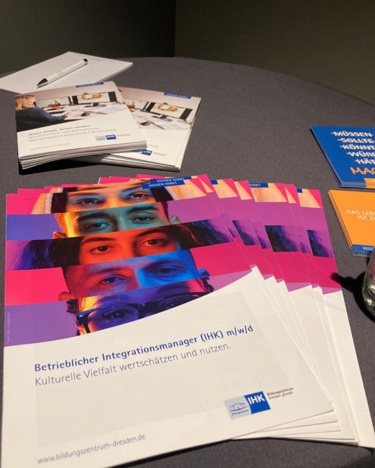 A stack of informational brochures is displayed on a table. The brochures feature diverse faces and highlight the role of an Integration Manager, emphasizing cultural diversity and its value. Nearby, there's a notepad and a pen, indicating an interactive event.