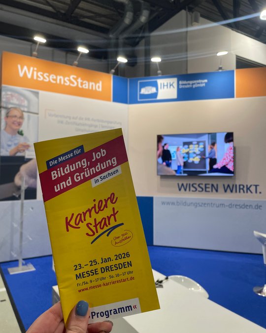 A person holds a yellow flyer for "Karriere Start," an event focused on education, jobs, and entrepreneurship in Saxony. The background features a booth with information about the event, including dates and a website link, emphasizing opportunities and resources available.