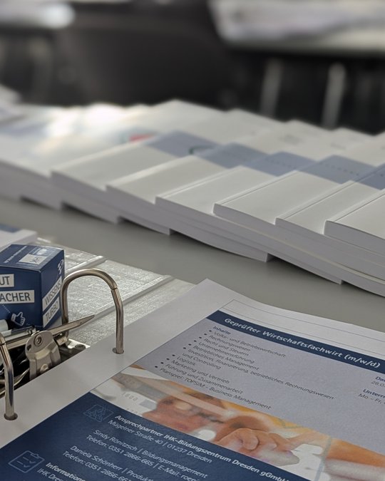 Auf dem Tisch liegen mehrere Stapel von Dokumenten und Broschüren. Im Vordergrund befindet sich ein Informationsblatt über die Ausbildung zum geprüften Wirtschaftsfachwirt (m/w/d) mit Stichpunkten zu Inhalten und Themen der Ausbildung. Ein gelochtes Blatt und ein kleiner Kasten sind ebenfalls sichtbar.