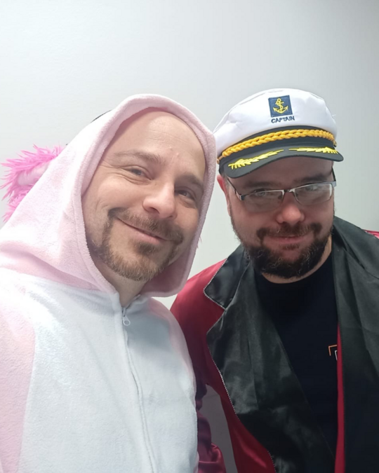 Two men pose for a selfie. One wears a pink unicorn onesie, complete with a horn and fluffy details, while the other is dressed in a captain's hat and a red jacket. They both smile, standing against a plain wall.