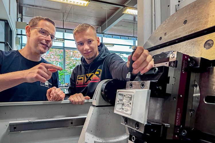 Two men are engaged in a conversation near a machine in a workshop. One man points towards the machine while the other holds a tool and looks at him attentively. Large windows in the background allow natural light into the workspace.
