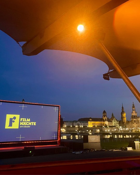Eine beleuchtete Leinwand mit dem Schriftzug "FILMNÄCHTE am Elbufer" steht im Vordergrund. Im Hintergrund sind teilweise erleuchtete historische Gebäude entlang des Flusses zu sehen, während der Himmel in der Dämmerung leicht erhellt ist.