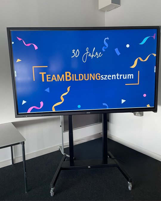 Ein Bildschirm zeigt den Schriftzug „30 Jahre TEAMBildungszentrum“ auf einem blauen Hintergrund, umgeben von bunten Feierdekorationen. Der Fokus liegt auf dem Jubiläum des Bildungszentrums.