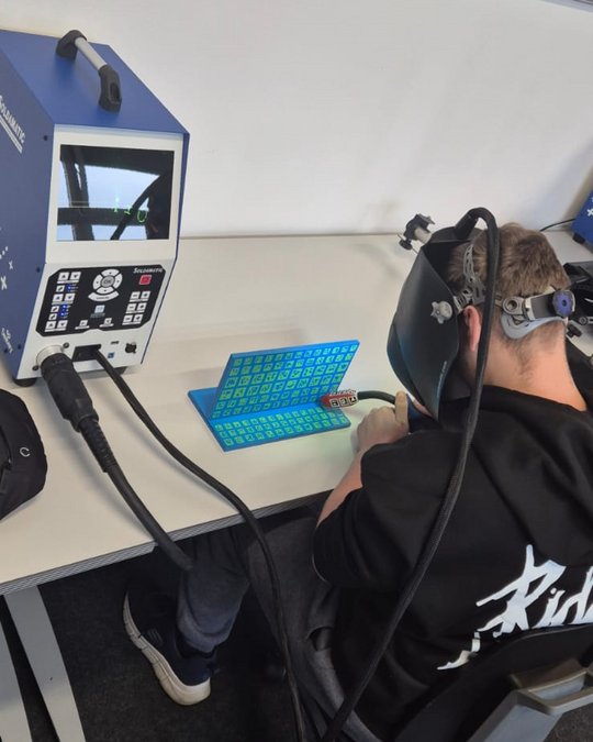 Eine Person sitzt an einem Tisch und arbeitet mit einem Schweißgerät, das an einer blauen Werkbank positioniert ist. Sie trägt eine Schweißmaske und hat ein Kabel in der Hand. Auf dem Tisch ist ein Bildschirm zu sehen, der möglicherweise Arbeitsanweisungen anzeigt.