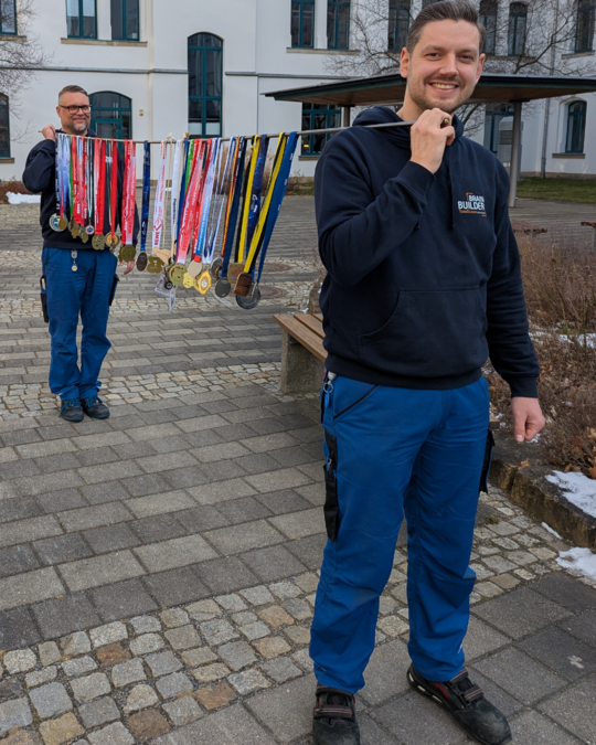 Two men stand outdoors, both wearing navy hoodies and blue pants. One man, in the foreground, smiles while holding a medal. The other man, in the background, showcases a collection of various medals displayed on a rack, highlighting their achievements.