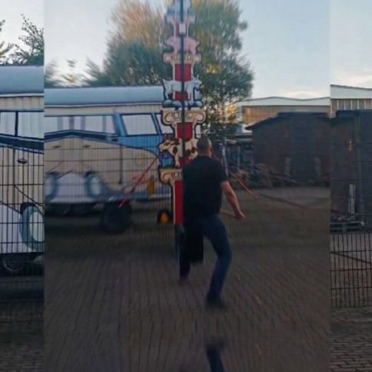 Eine Person steht vor einem dekorierten Pfahl in einem Außenbereich. Im Hintergrund sind Wohnmobile und eine geschlossene Umgebung sichtbar. Die Person hat einen Rucksack und scheint sich in Bewegung zu befinden. Der Himmel ist leicht bewölkt.