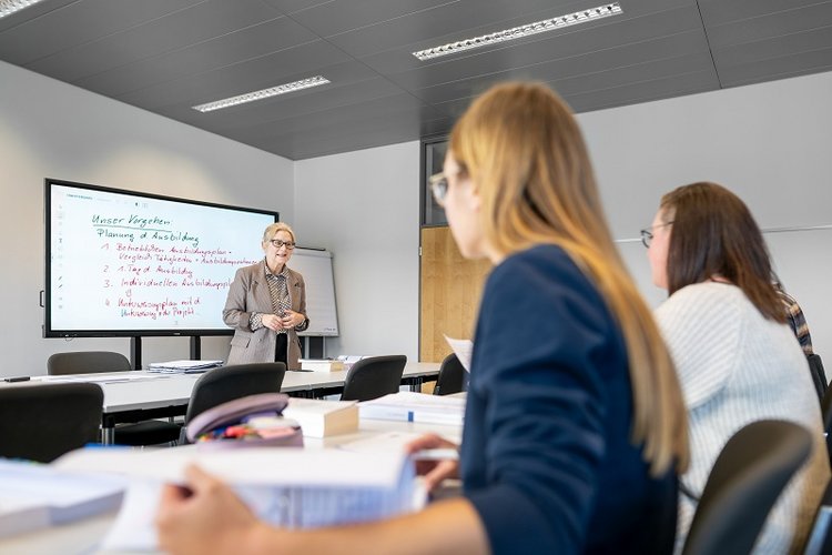 A teacher stands in front of a whiteboard, explaining a lesson to students seated at desks. The classroom has an organized arrangement with books and materials on the tables. The atmosphere is focused on learning and engagement.