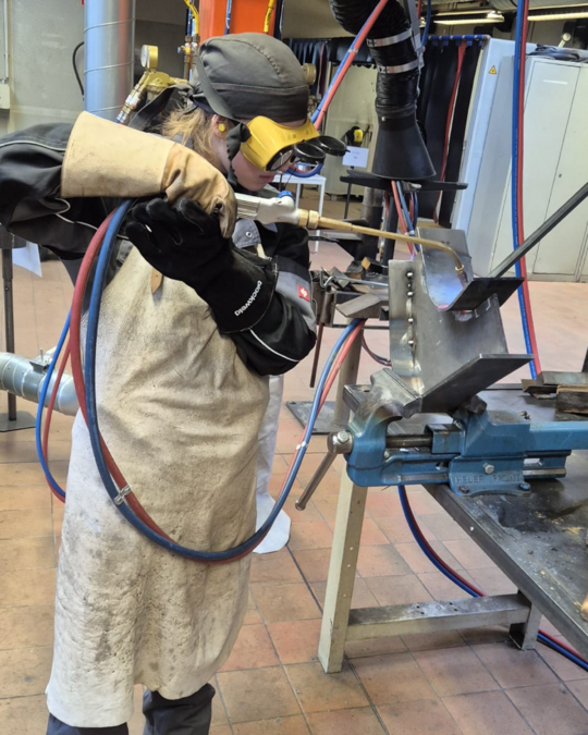 A person in protective gear, including a helmet and gloves, is using a welding torch to work on a metal piece secured in a vise. The setting appears to be a workshop with equipment in the background.