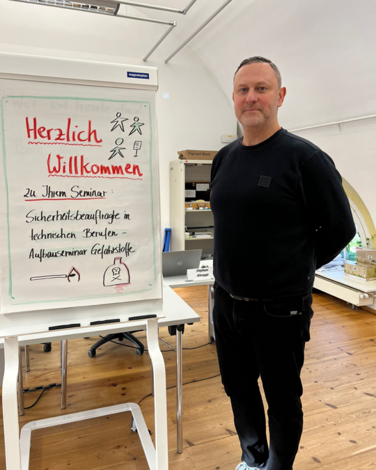 A man stands beside a whiteboard that reads "Herzlich Willkommen" and details about a seminar on safety protocols in technical professions. The setting appears to be an indoor conference space with wooden flooring. The man is wearing a black sweater and has a welcoming expression.