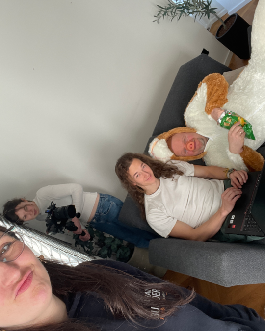 A group of four people is gathered in a cozy living room. One person is taking a selfie, while another is sitting on a couch with a laptop, and a third is standing behind holding a camera. A large plush toy is next to the seated person.