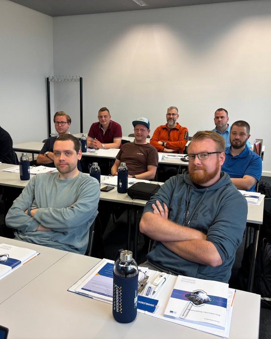 A group of men sits in a classroom setting, attentively facing forward. They are arranged in rows at tables with papers and water bottles in front of them. The atmosphere appears focused and engaged.