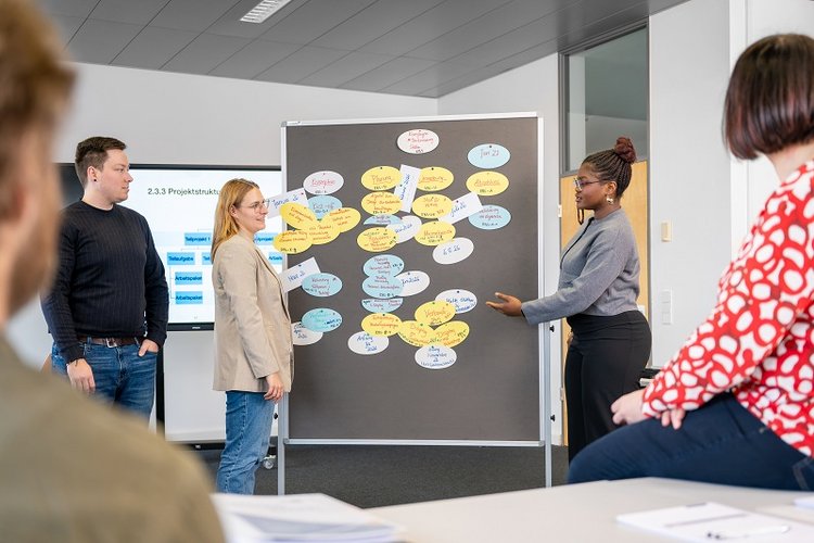 A group of four people engages in a collaborative discussion around a presentation board filled with colorful notes. One person is presenting ideas, while others listen attentively, suggesting a dynamic brainstorming session in a professional setting.