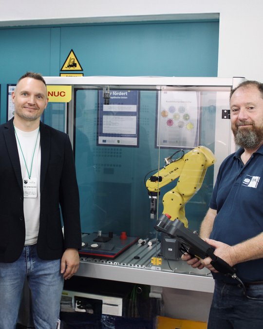 Zwei Männer stehen neben einem automatisierten Roboterarm in einem Ausbildungsraum. Der linke Mann trägt einen Blazer und hat ein Namensschild, während der rechte Mann ein T-Shirt trägt und ein Werkzeug in der Hand hält. Im Hintergrund sind verschiedene Bedienfelder sichtbar.