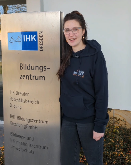 A person wearing glasses stands next to a metal sign displaying "Bildungszentrum" and the logo for IHK Dresden. The individual is smiling and wearing a dark hoodie. The background suggests an outdoor setting near the educational institution.