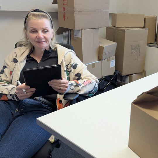 A woman with long hair, wearing a patterned cardigan and black top, sits in an office chair while holding a tablet. She appears to be engaged with the device. In the background, there are stacked cardboard boxes, indicating a storage or unpacking scenario.