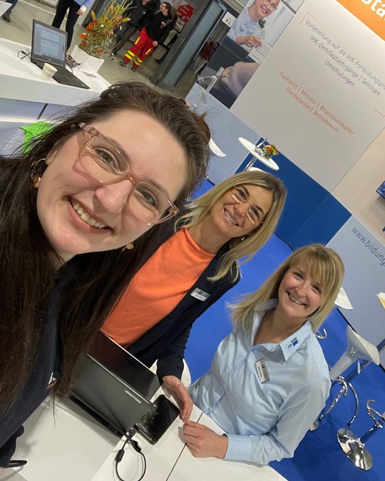 Three women are smiling and posing for a selfie at a trade show or conference booth. They are wearing professional attire and standing in front of a display with promotional materials. The atmosphere appears friendly and inviting.