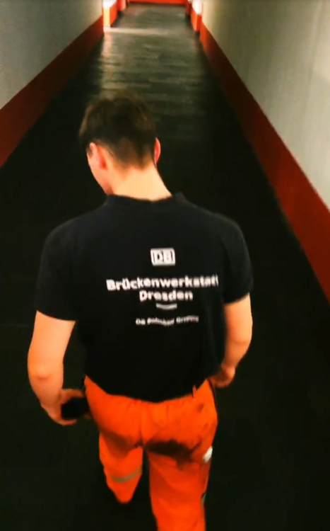A person wearing an orange work uniform walks down a dimly lit corridor. The back of their navy shirt displays "Brückenwerkstatt Dresden" and a logo. The corridor has red and white walls, leading to a brightly lit area ahead.
