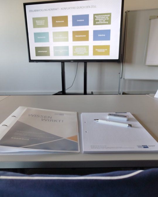 A presentation screen displays various colored blocks with text. In the foreground, a folder labeled "WISSEN WIRKT!" is placed on a table alongside blank papers and a pen. The setting appears to be a professional meeting or training room.