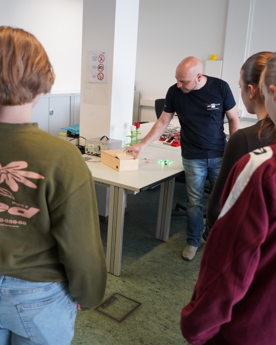A man demonstrates a small project on a table, with several students observing. The table has various tools and materials, including a wooden box and green lights. The setting appears to be a classroom or workshop.