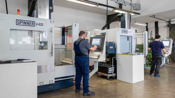 Two workers are operating CNC machines in a manufacturing facility. One worker stands in front of a control panel on the left, focusing on the machine's display, while another worker is at a different machine on the right, also engaged with a control panel.