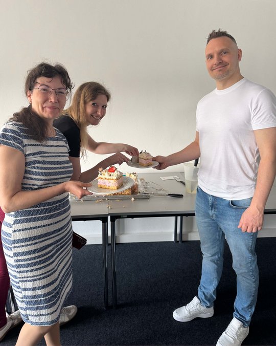 Drei Personen stehen um einen Tisch mit einem Kuchenteller. Zwei Frauen schneiden ein Stück Kuchen an, während ein Mann neben ihnen lächelt. Alle scheinen Freude zu haben und die Atmosphäre wirkt freundlich und festlich.