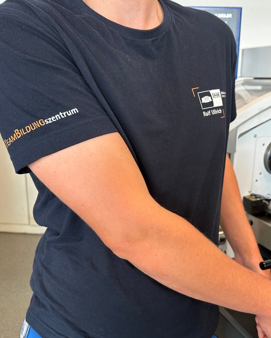 Ein Teil eines Armes in einem dunkelblauen T-Shirt ist sichtbar. Auf dem Ärmel steht "TeamBILDUNGSzentrum". Im Hintergrund sind Maschinen und ein heller Raum zu erkennen, jedoch nicht im Fokus des Bildes.