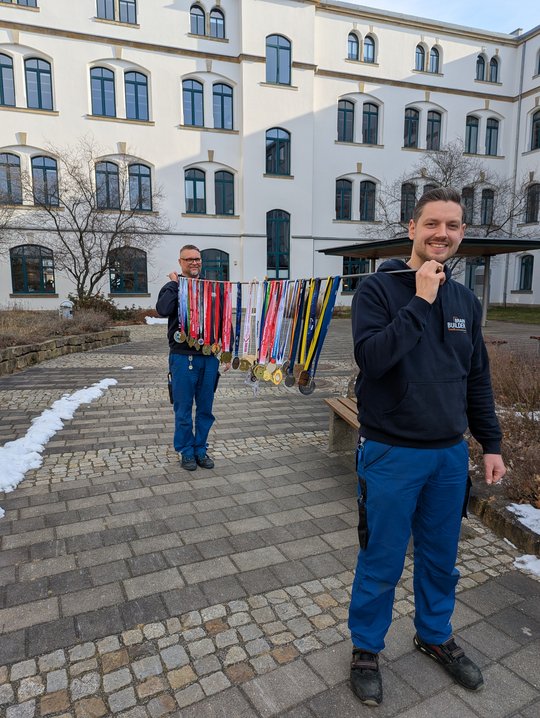 Two men are standing outside, one in front of the other, both dressed in blue work pants and dark tops. The man in the foreground smiles while holding a display of colorful medals, showcasing achievements. A building with multiple windows is visible in the background.