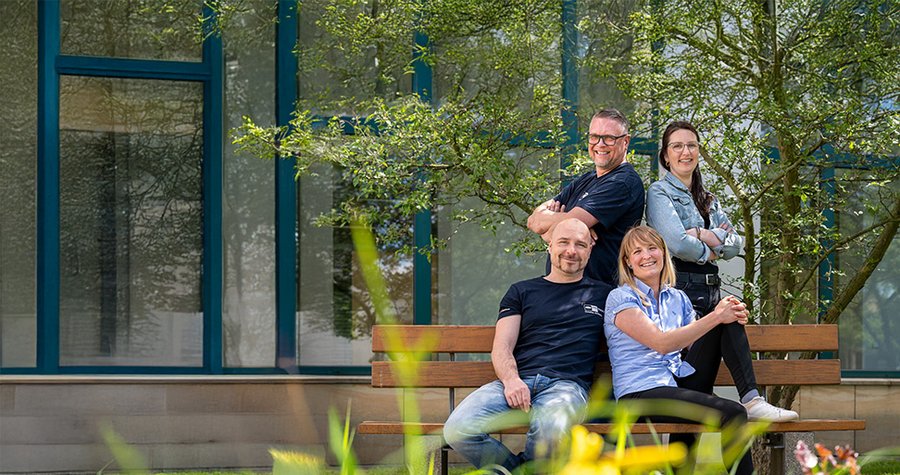 Four people are sitting on a bench in a sunny outdoor setting, with greenery in the background. Two men and two women are smiling and posing, creating a friendly and relaxed atmosphere. The scene conveys a sense of teamwork and camaraderie.