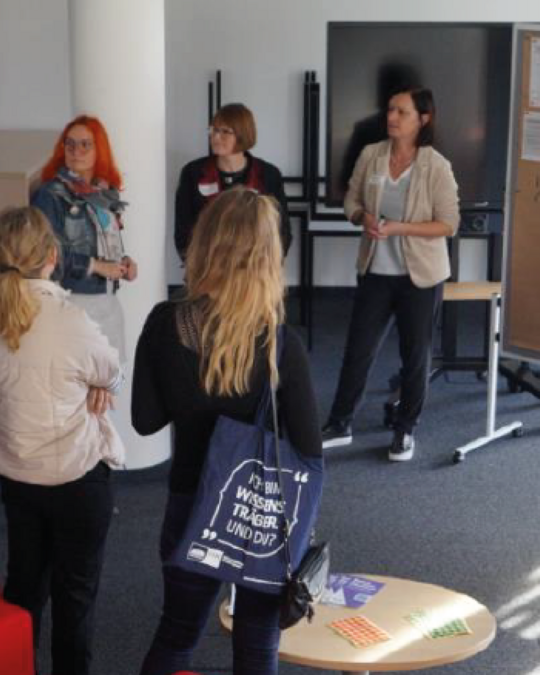 A group of five people is gathered in a room for a discussion. Three women stand facing the group, while two individuals with their backs to the camera observe. One woman has bright orange hair and wears glasses, while another holds a notepad. A small round table displays various materials.