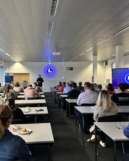 A speaker stands in front of an audience in a conference room, addressing them about a topic. The audience, consisting of various individuals seated at tables, faces a screen displaying a logo. The environment is well-lit and organized for a professional presentation.