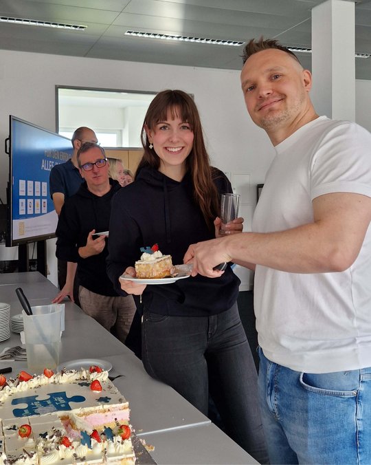 Zwei Personen stehen an einem Tisch mit einer Torte. Eine Frau hält einen Teller mit einem Stück Torte, während ein Mann ein weiteres Stück serviert. Im Hintergrund sind weitere Personen zu sehen, die an der Feier teilnehmen.
