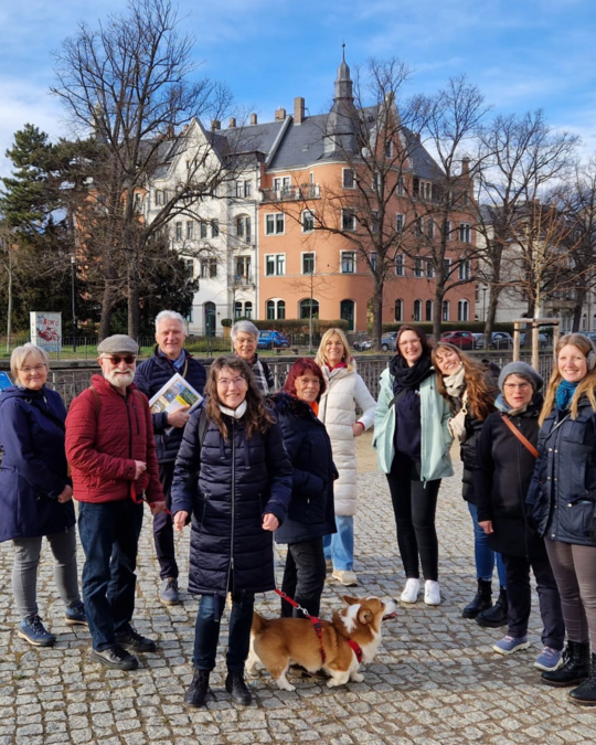 A group of diverse people, including men and women of varying ages, gather outdoors on a cobblestone path, with buildings and trees in the background. A corgi dog stands in front. Everyone is smiling and engaging with one another.