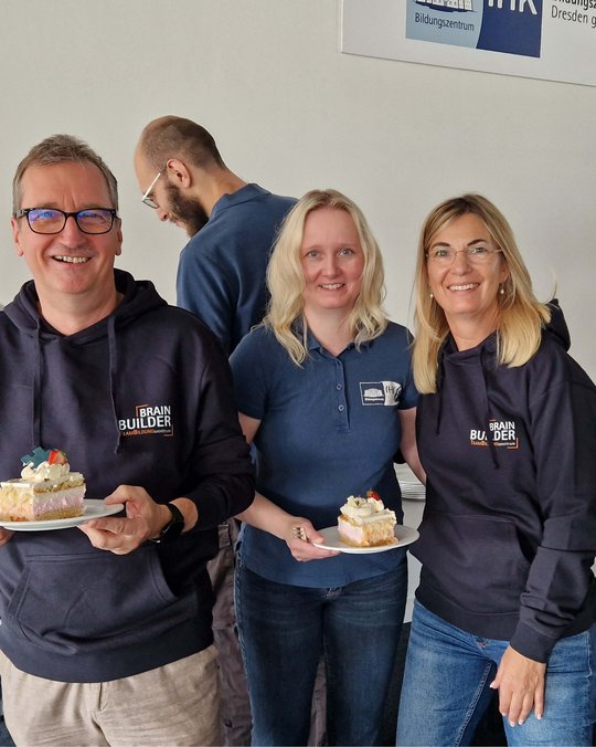 Drei Personen in Sweatshirts mit dem Aufdruck "BRAIN BUILDER" halten lächelnd Kuchenstücke auf Tellern. Sie stehen in einer freundlichen Atmosphäre, während eine weitere Person im Hintergrund zu sehen ist.
