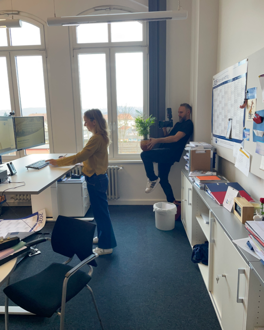 A woman sits at a desk, typing on a keyboard, while a man leans against a wall nearby, holding a potted plant. Both are in a well-lit office space with windows and office furniture in the background.