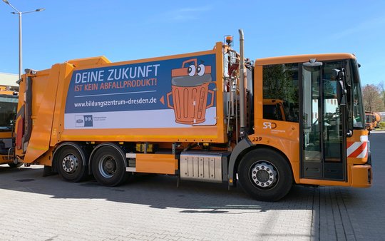 A bright orange garbage truck is parked, displaying a large advertisement on its side. The ad promotes the message "Your future is not waste!" along with a website link. The background features a clear blue sky and a paved area.