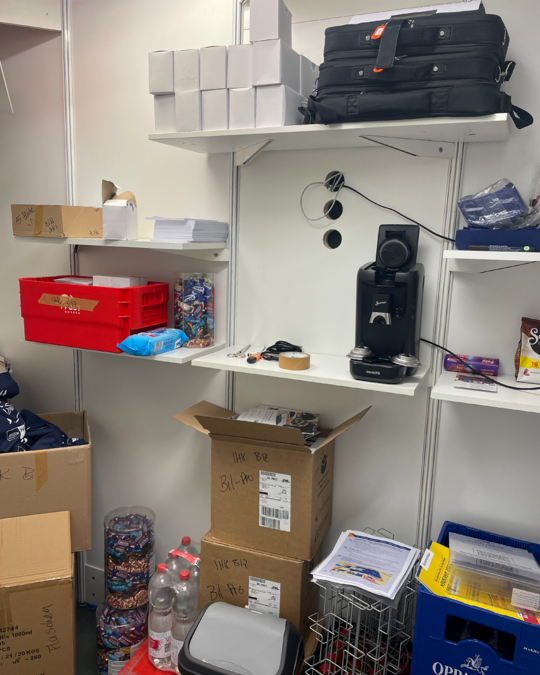 A cluttered storage area featuring shelves with boxes, a small black suitcase, and a coffee machine. Various cardboard boxes and containers are stacked below, along with some water bottles. The scene suggests a workspace or storage setup with organizational items.