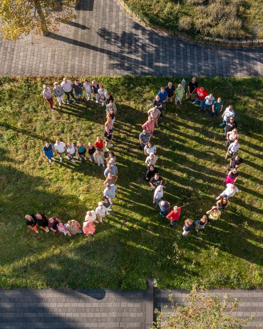 Eine Gruppe von Menschen steht auf einer Wiese und bildet die Zahl "30". Die Szene wird aus der Vogelperspektive aufgenommen, und die Menschen erzeugen durch ihre Anordnung Schatten auf dem Boden. Es gibt einen klaren Himmel und teilweise Schatten durch Bäume.