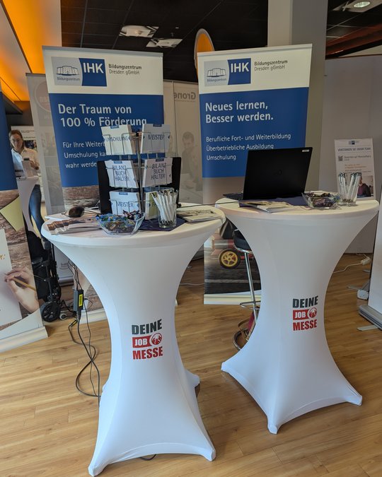 Zwei Stehtische auf einer Messe mit Informationsmaterialien über berufliche Weiterbildung und Umschulung. Im Hintergrund stehen Banner des Bildungszentrums Dresden, die Themen wie Förderung und Verbesserung durch Lernen ansprechen. Ein Laptop und Schreibutensilien sind ebenfalls auf den Tischen platziert.