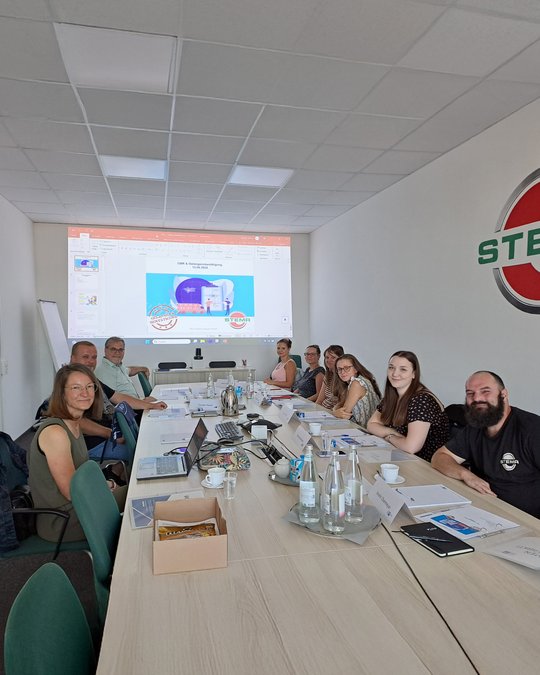 Eine Gruppe von sieben Personen sitzt an einem Konferenztisch in einem hellen Raum. Auf der Wand projiziert eine Präsentation mit Grafiken und Text. Einige Notebooks, Wasserflaschen und Unterlagen sind auf dem Tisch zu sehen, während die Teilnehmer aufmerksam dem Vortrag folgen.