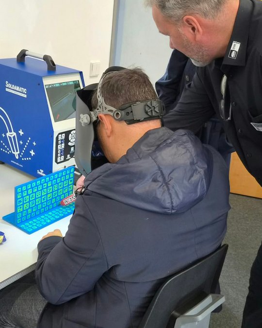 A man wearing a welding helmet is seated at a table, using a colorful keyboard to interact with a welding simulation machine. Another individual, standing behind him, is observing his actions. The setting appears to be an educational or training environment.