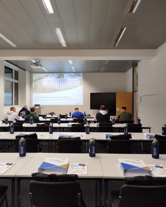 A classroom setting is shown with several rows of desks. Participants are seated, facing a presentation screen displaying a welcome message in German. Each desk has water bottles and course materials, indicating an educational event or workshop.